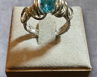 Puede incluir: Un anillo de plata con alambre envuelto con una piedra preciosa cuadrada de color azul claro. La banda del anillo está intrincadamente tejida, y la piedra preciosa se sujeta con alambres enrollados. El anillo se exhibe sobre un soporte beige cubierto de tela.