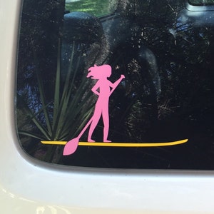 May include: Pink silhouette of a person paddleboarding on a yellow surfboard. The sticker is on a car window.