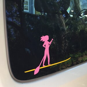 May include: Pink silhouette of a person paddleboarding on a yellow paddleboard. The sticker is on a car window.