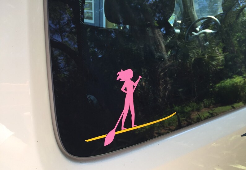 May include: Pink silhouette of a person paddleboarding on a yellow paddleboard. The sticker is on a car window.