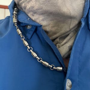 May include: A silver chain necklace with cylindrical links. The necklace is worn by a person wearing a blue shirt.