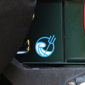 May include: A blue and white sticker with a wave and trident design is on the back of a green car. The sticker is in a circle.
