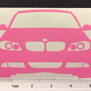 May include: A pink car decal featuring a front view of a car. The decal is a solid pink color and has a simple, stylized design. The image includes a ruler with measurements in inches.