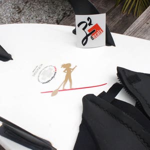 May include: A white stand up paddleboard with black fins and a black wetsuit. The paddleboard has a red line and a sticker with a woman paddling and the text "SurftoFeet". There is another sticker with the text "J2 Decals".
