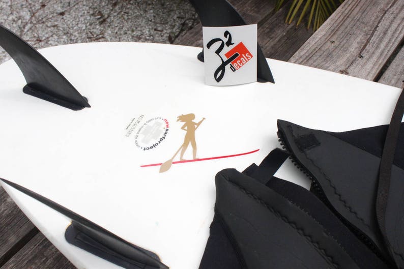 May include: A white stand up paddleboard with black fins and a black wetsuit. The paddleboard has a red line and a sticker with a woman paddling and the text "SurftoFeet". There is another sticker with the text "J2 Decals".