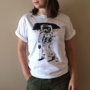 May include: A white t-shirt with a black and white image of an astronaut on the moon. The astronaut is wearing a spacesuit and a helmet. The t-shirt has rolled up sleeves and a crew neck. The person wearing the t-shirt is also wearing green cargo shorts.