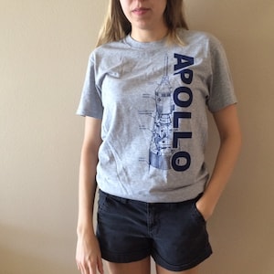 May include: A gray t-shirt with a blue and white illustration of a rocket ship and the word "APOLLO" printed vertically on the right side of the shirt.