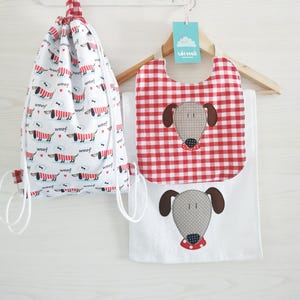 Può includere: Un set bianco composto da zaino con coulisse, bavaglino e asciugamano con un design a forma di cane bassotto. Lo zaino e il bavaglino hanno un motivo a quadri rossi e bianchi. Il cane ha orecchie marroni e un colletto rosso. La parola "woof" è stampata.