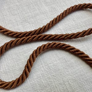 May include: A brown twisted rope cord featuring a thick, multi-strand construction with a smooth, satin-like sheen. The cord is arranged in a loose, flowing S-shape, showcasing the uniform spiral texture and consistent dark brown color throughout.