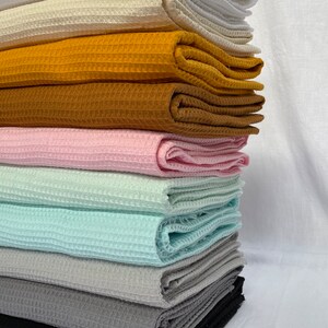 Puede incluir: Una pila de ocho muestras de tejido de tejido de waffle en varios colores, incluyendo blanco, crema, amarillo mostaza, marr&oacute;n, rosa, azul claro, gris y negro.