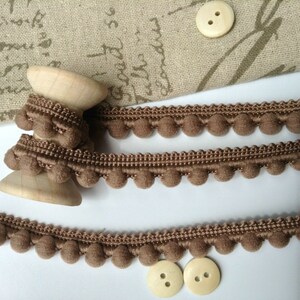 May include: Brown pom-pom trim with a wooden spool. The trim is made of soft, fuzzy pom-poms and a woven base. Two beige buttons are also shown.