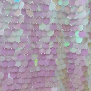 Op de afbeelding: Close-up van een stof bedekt met iriserende pailletten. De pailletten zijn rond en gerangschikt in een herhalend patroon. De kleuren verschuiven van lichtroze naar wit en groen.