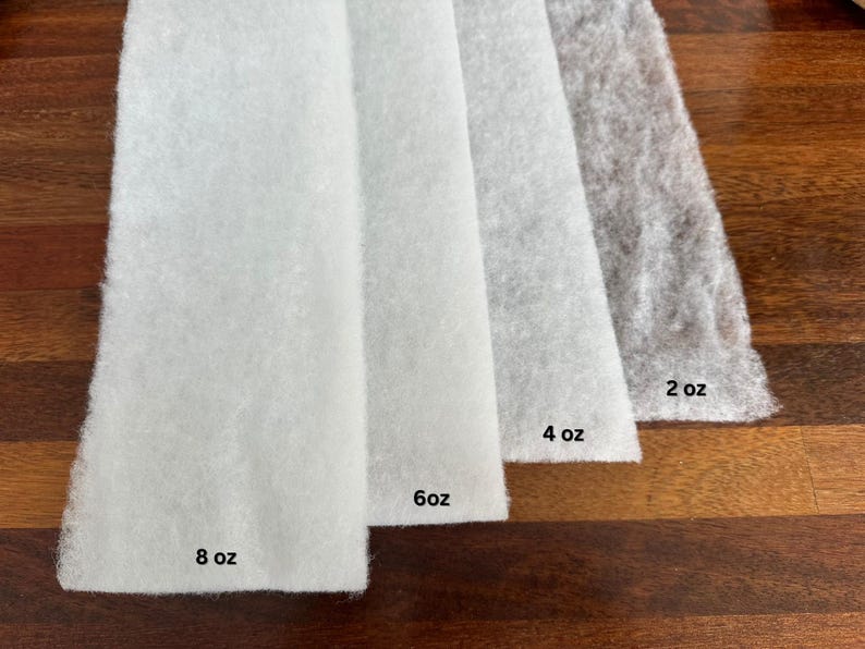 May include: Four rectangular pieces of white and gray batting are displayed on a wooden surface. The batting pieces are labeled with their weights: 8 oz, 6 oz, 4 oz, and 2 oz. The batting has a soft, fluffy texture.