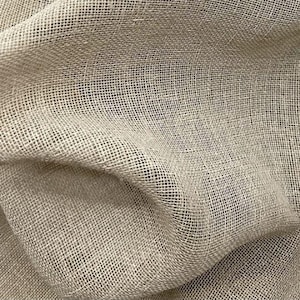 Può includere: Primo piano di un tessuto beige, a trama larga, con un motivo strutturato a griglia. Il tessuto sembra essere drappeggiato, con pieghe morbide e ombre che aggiungono profondità. Il materiale ha un aspetto naturale e organico.
