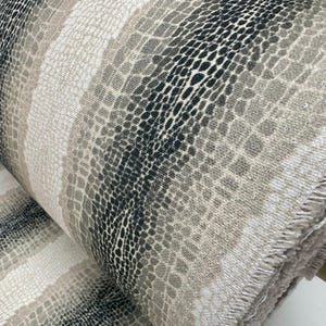 May include: A roll of fabric with a snakeskin design. The material displays a repeating scale pattern in grey, black, and beige tones. The pattern is arranged in vertical stripes, giving a textured look. Suitable for upholstery or fashion applications.