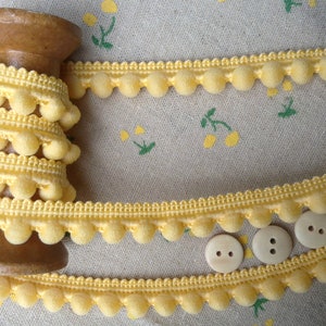 May include: A spool of yellow pom-pom trim with a wooden spool. The trim is a single row of pom-poms with a thin, woven base.