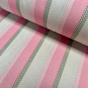 May include: Close-up of a fabric roll featuring a repeating pattern of vertical stripes. The stripes alternate between light pink, off-white, and a light green herringbone design. The fabric appears to be cotton or a similar material.