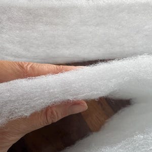 May include: Close-up of a handful of fluffy, white polyester fiberfill. The soft, cloud-like material is held in a hand, with more of the filling visible in the background. The texture is fine and airy, suitable for stuffing.