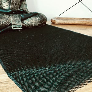 May include: A dark green woven yoga mat with a textured surface and fringed edges. A seated Buddha statue and a wooden incense holder are in the background, suggesting a focus on meditation and relaxation.