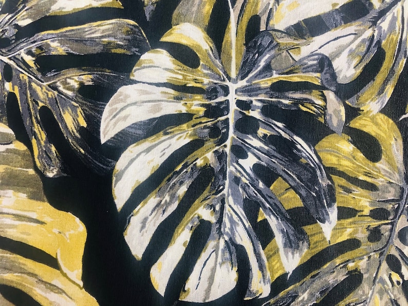 May include: A black fabric with a large white and yellow leaf pattern. The leaf has a detailed vein pattern and is reminiscent of a monstera plant.