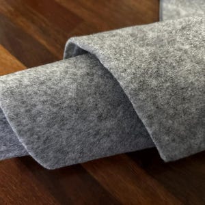 May include: Close-up of folded grey felt fabric. The felt has a soft, textured appearance and is layered to reveal its thickness. The background is a dark wooden surface.