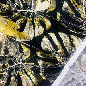 May include: Black fabric with a large-scale tropical leaf print in shades of gold, beige, and gray. The fabric is shown folded over, revealing the white selvedge edge.