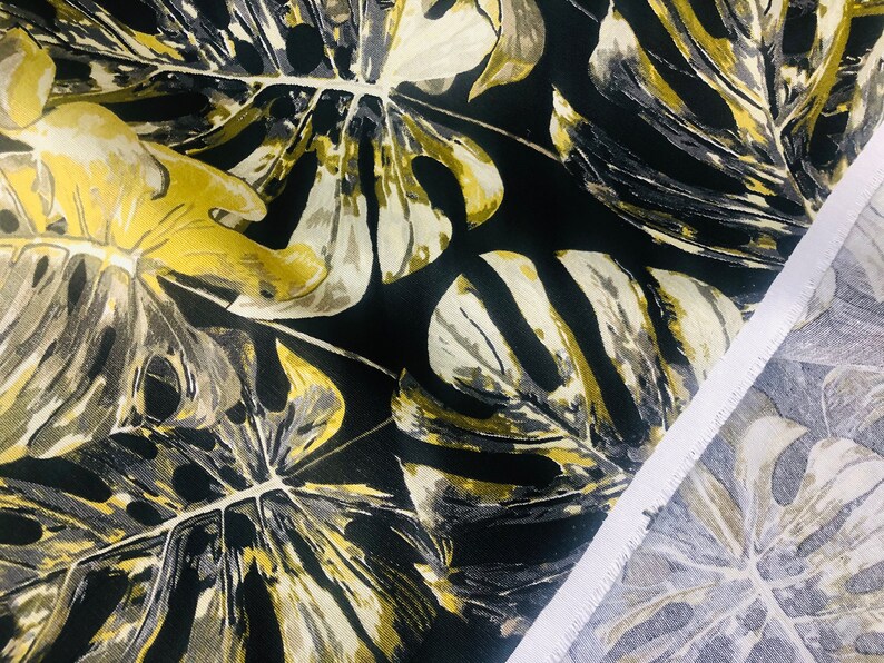 May include: Black fabric with a large-scale tropical leaf print in shades of gold, beige, and gray. The fabric is shown folded over, revealing the white selvedge edge.