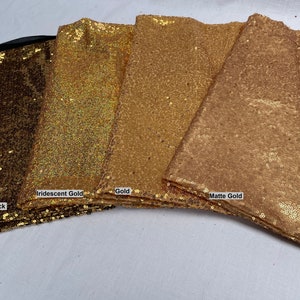 Pode incluir: Close-up de quatro tecidos de lantejoulas douradas diferentes. Os tecidos s&atilde;o rotulados como "Gold on Black", "Iridescent Gold", "Gold" e "Matte Gold". Os tecidos est&atilde;o dobrados e empilhados, mostrando suas diferentes texturas e cores.