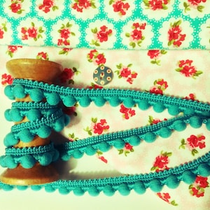 May include: Teal pom-pom trim wound on a wooden spool, against a floral fabric backdrop. The trim is a decorative embellishment for sewing and crafting projects.