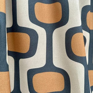 Op de afbeelding: Een stof met een herhalend geometrisch patroon van afgeronde rechthoekige vormen in bruin en donker antracietgrijs, geplaatst tegen een lichtcr&egrave;me achtergrond. Het textiel vertoont een consistent, gestructureerd ontwerp met zachte plooien.