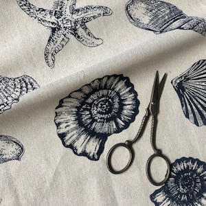 May include: A white fabric with a navy blue print of seashells and starfish. A pair of antique scissors are lying on the fabric.