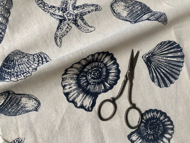 May include: A white fabric with a navy blue print of seashells and starfish. A pair of antique scissors are lying on the fabric.