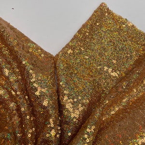 Pode incluir: Close-up de tecido dourado com lantejoulas. O tecido est&aacute; dobrado, revelando um padr&atilde;o em forma de V. As lantejoulas s&atilde;o pequenas, redondas e iridescentes, refletindo a luz. A cor geral &eacute; um dourado quente com toques de outras cores.