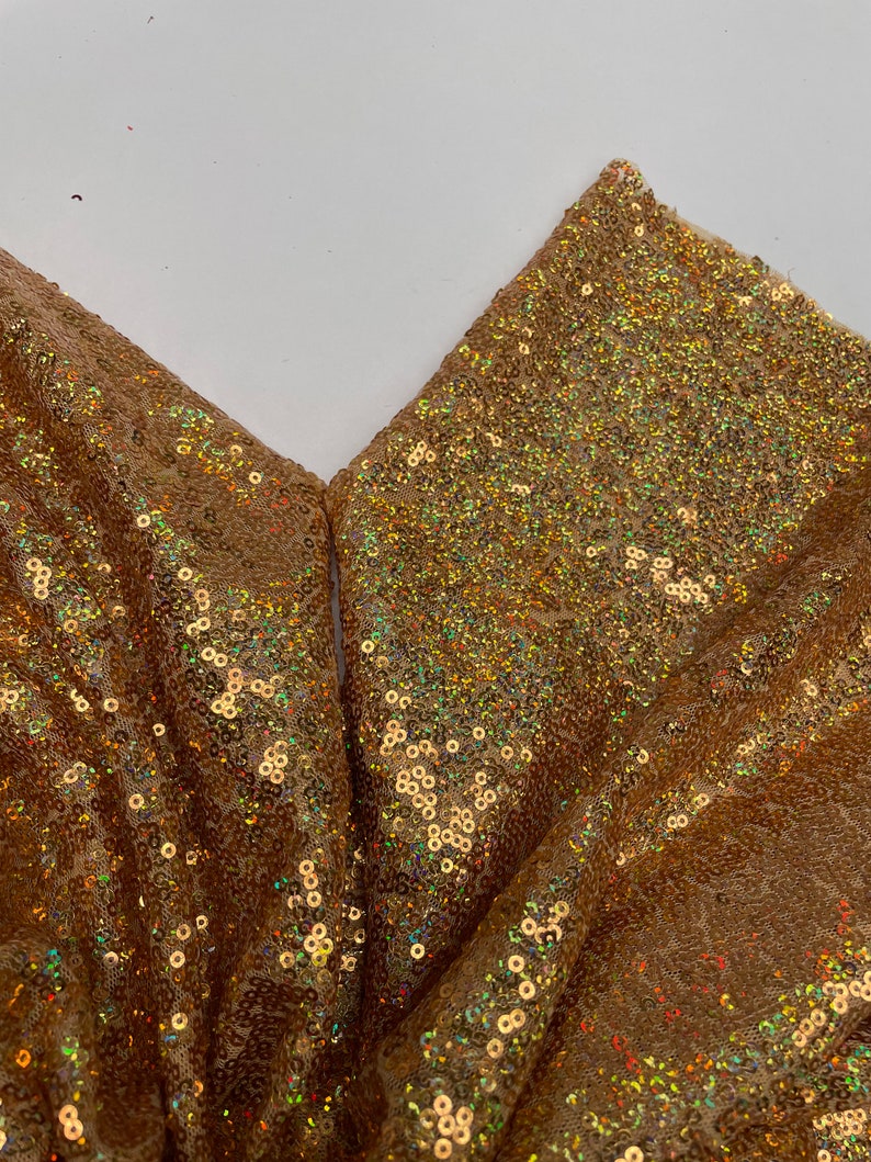 Pode incluir: Close-up de tecido dourado com lantejoulas. O tecido est&aacute; dobrado, revelando um padr&atilde;o em forma de V. As lantejoulas s&atilde;o pequenas, redondas e iridescentes, refletindo a luz. A cor geral &eacute; um dourado quente com toques de outras cores.