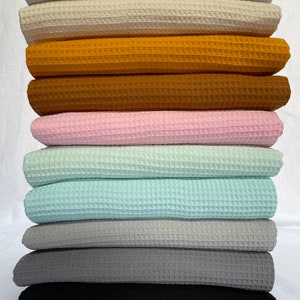 Puede incluir: Una pila de tela de tejido de waffle en varios colores, incluyendo blanco, beige, amarillo mostaza, marr&oacute;n, rosa, azul claro, gris y negro.