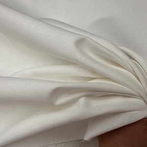 Puede incluir: Tela blanca con una superficie lisa y ligeramente texturizada. La tela está doblada y drapeada, mostrando su textura suave y fluida.