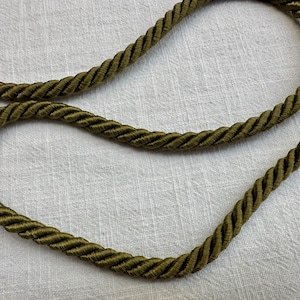 May include: A single length of olive green twisted cord trim featuring a thick, rope-like construction with a smooth, satin-like sheen. The cord is arranged in a loose, curved loop, showcasing the uniform spiral texture of the twisted fibers.