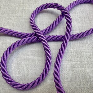 May include: A length of vibrant purple twisted cord featuring a smooth, glossy finish and a multi-strand braided construction. The cord is arranged in a loose, overlapping loop pattern, showcasing the consistent texture and deep violet color.