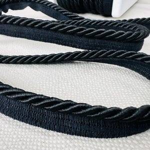 Puede incluir: Borde trenzado negro con una textura gruesa y tejida. El borde está hecho de un material suave y flexible y tiene un acabado liso y pulido.