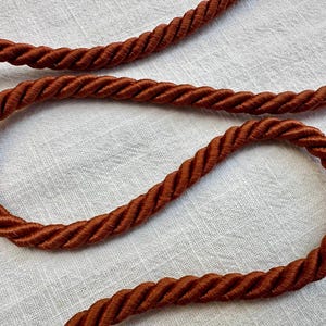 May include: A thick, rust-colored twisted cord made of multiple strands of fiber wound together. The cord features a distinct spiral texture and a deep, warm brown-orange hue, showing a consistent rope-like construction throughout its length.