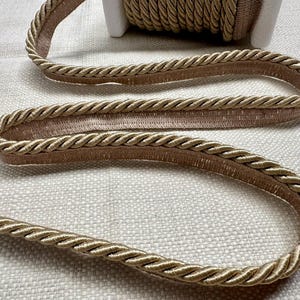 May include: A close-up of a beige braided trim with a subtle shimmer. The trim is thick and has a rope-like texture. It is perfect for adding a touch of elegance to any project.