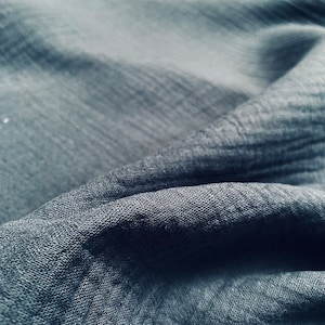 May include: Close-up of a dark gray fabric with a textured weave. The fabric is folded and draped, creating soft folds and shadows.