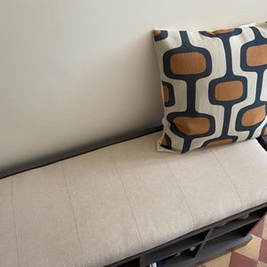 May include: A rectangular storage bench with a dark frame and a light beige upholstered seat cushion. A square throw pillow rests on the seat, featuring a repeating pattern of dark gray rounded rectangular shapes and solid brown oval centers.
