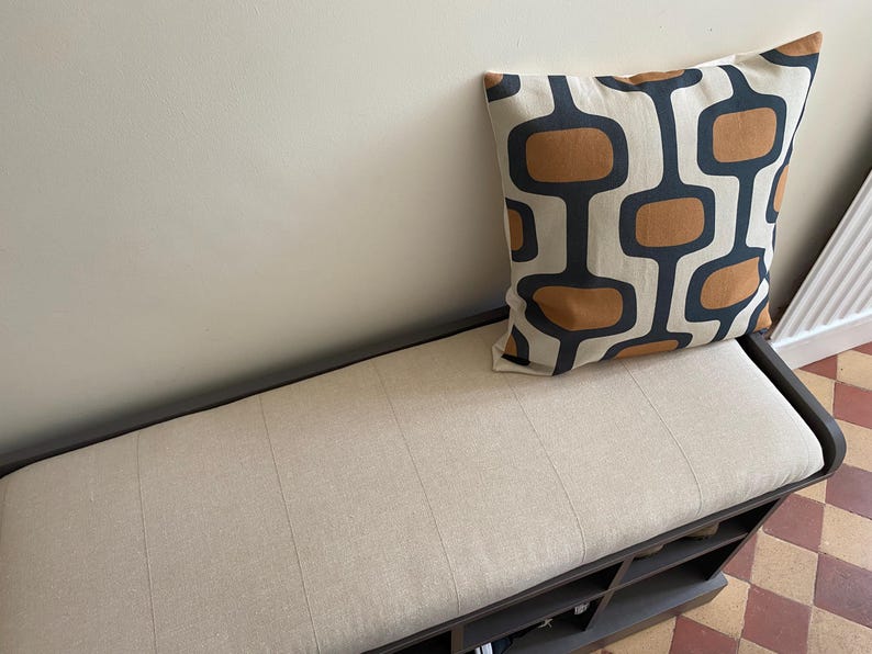 May include: A rectangular storage bench with a dark frame and a light beige upholstered seat cushion. A square throw pillow rests on the seat, featuring a repeating pattern of dark gray rounded rectangular shapes and solid brown oval centers.