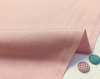 Puede incluir: Primer plano de una tela rosa pálido con una textura sutil. La tela está doblada por la mitad y se ven dos pequeños botones con lunares en la esquina inferior derecha.