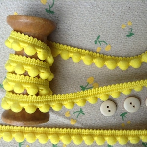 May include: A wooden spool with bright yellow pom-pom trim wound around it, alongside three small, round, off-white buttons. The trim features a scalloped edge and evenly spaced pom-poms. The background is a light fabric with a yellow floral pattern.