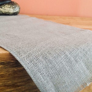 Peut inclure: Un chemin de table en toile de jute gris clair avec une texture tissée. Le chemin de table est drapé sur une surface en bois, avec une statue de Bouddha en arrière-plan. Les bords du chemin de table sont effilochés.