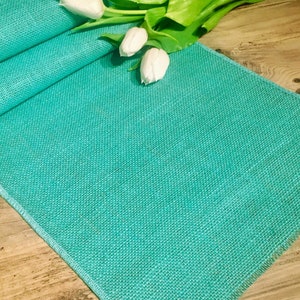 May include: A teal burlap table runner with a frayed edge. The runner is folded in half and a few white tulips are laying on top of it.