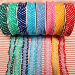 Op de afbeelding: Een verzameling spoelen met stippenlint in verschillende kleuren, waaronder groen, turquoise, marineblauw, lichtblauw, roze, geel en rood. Elke spoel heeft een witte rand en een bijpassend lint dat naar beneden loopt. De linten worden gepresenteerd op een rood-wit gestreept oppervlak.