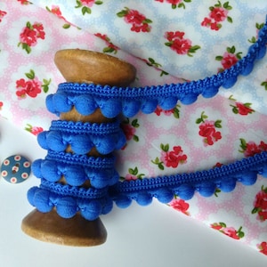 May include: A spool of bright blue pom-pom trim with a wooden bobbin. The trim is on top of a pink and blue floral fabric.